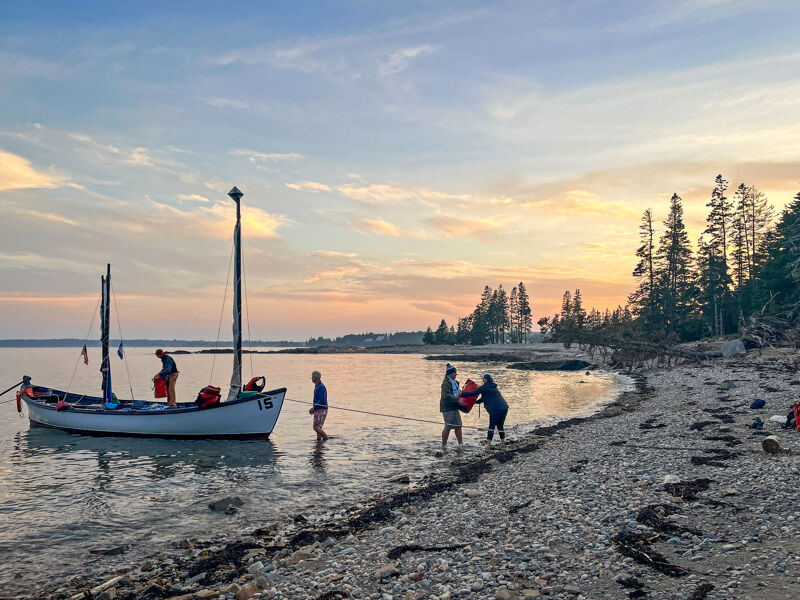 The image shows a serene coastal scene at sunset or sunrise. A small sailboat is anchored near the shore, with people standing on the beach and in the shallow water. The sky is filled with soft, warm colors, and the calm water reflects the light. The beach is rocky, and trees line the coast in the background, creating a peaceful and picturesque setting.